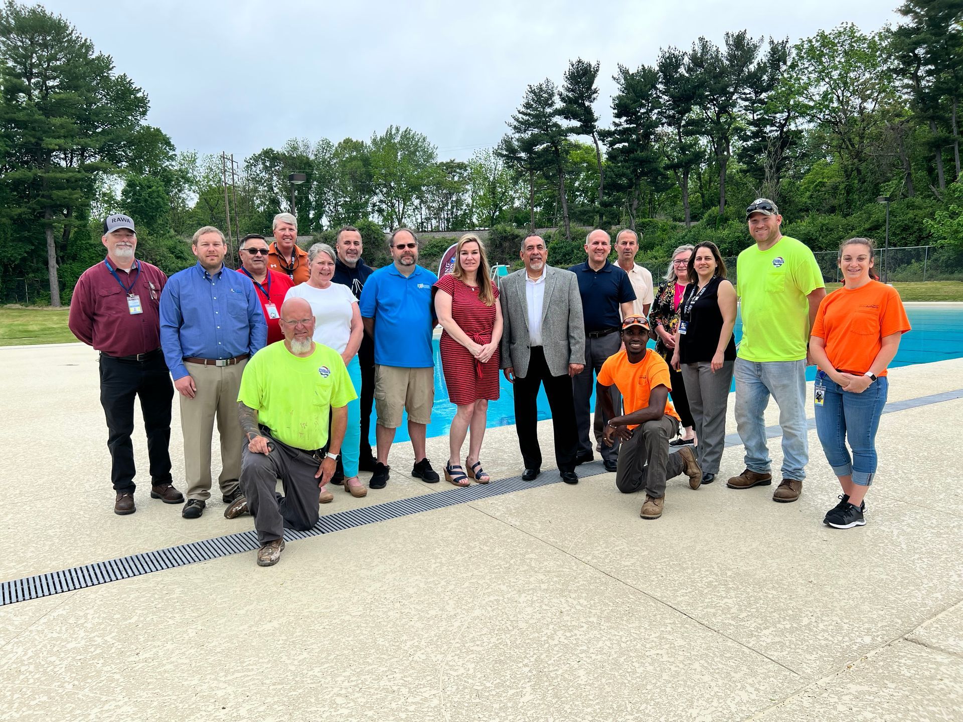 Group of people standing in front of a pool. People wearing casual attire. Trees in the background.