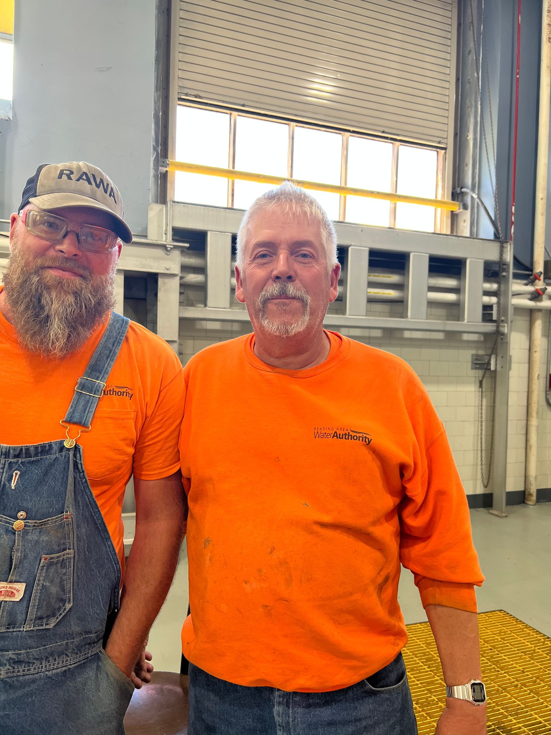 Two men in orange shirts, one with overalls and a hat, stand together. Industrial setting.