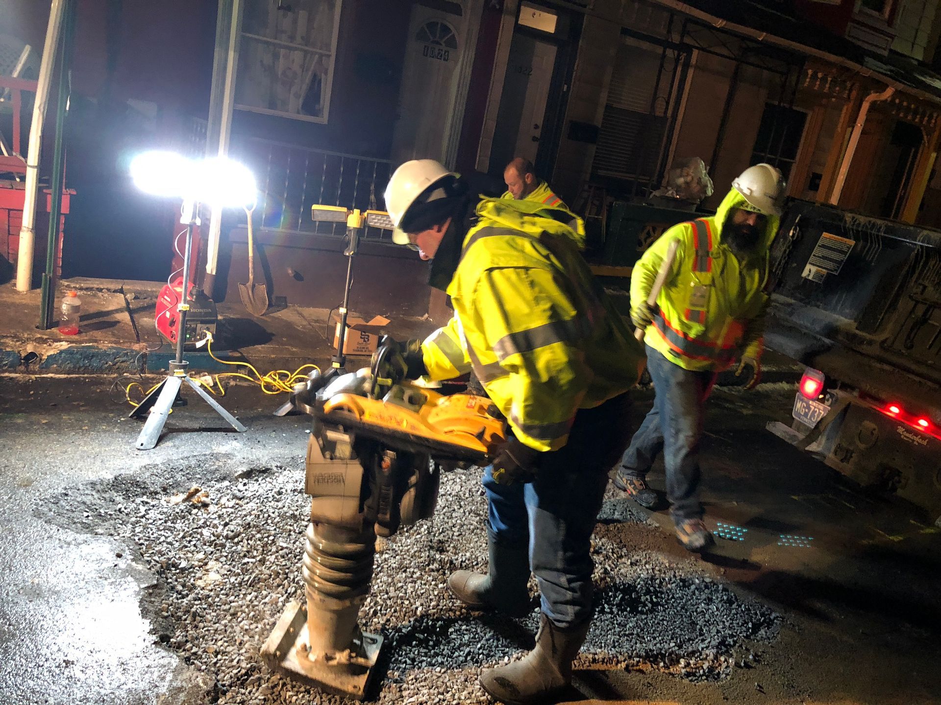 Two workers in reflective vests compacting asphalt at night with a machine under a bright light.