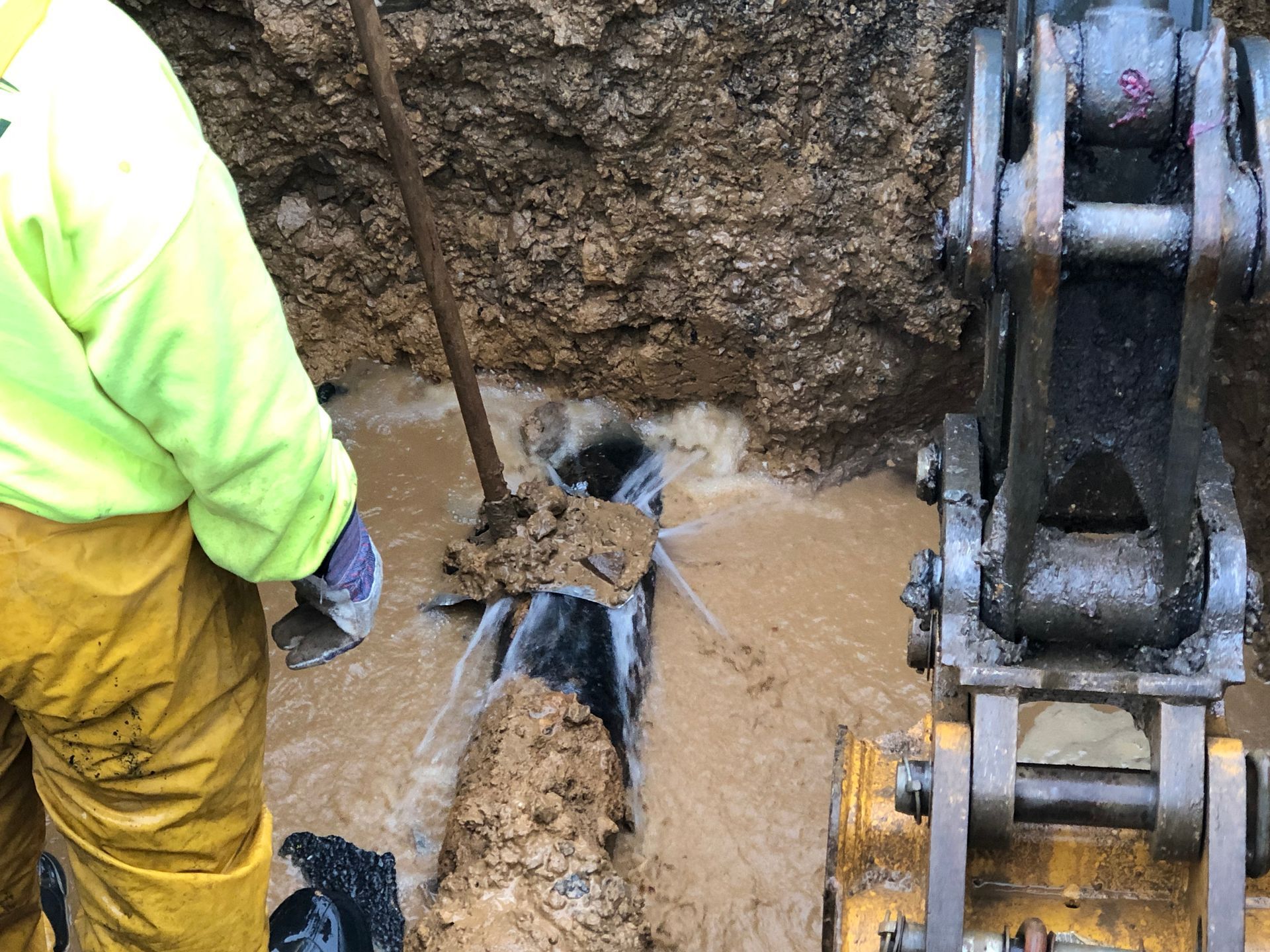 Worker in yellow rain gear near exposed pipe gushing water, with an excavator arm.