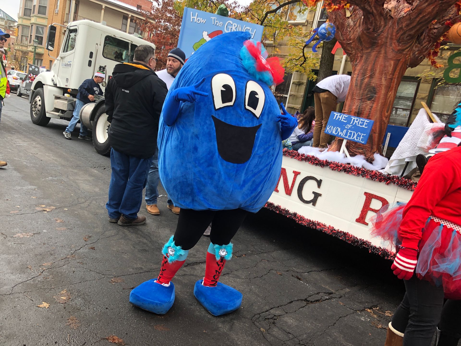 Blue mascot in costume at parade, smiling with arms out. Float in background.