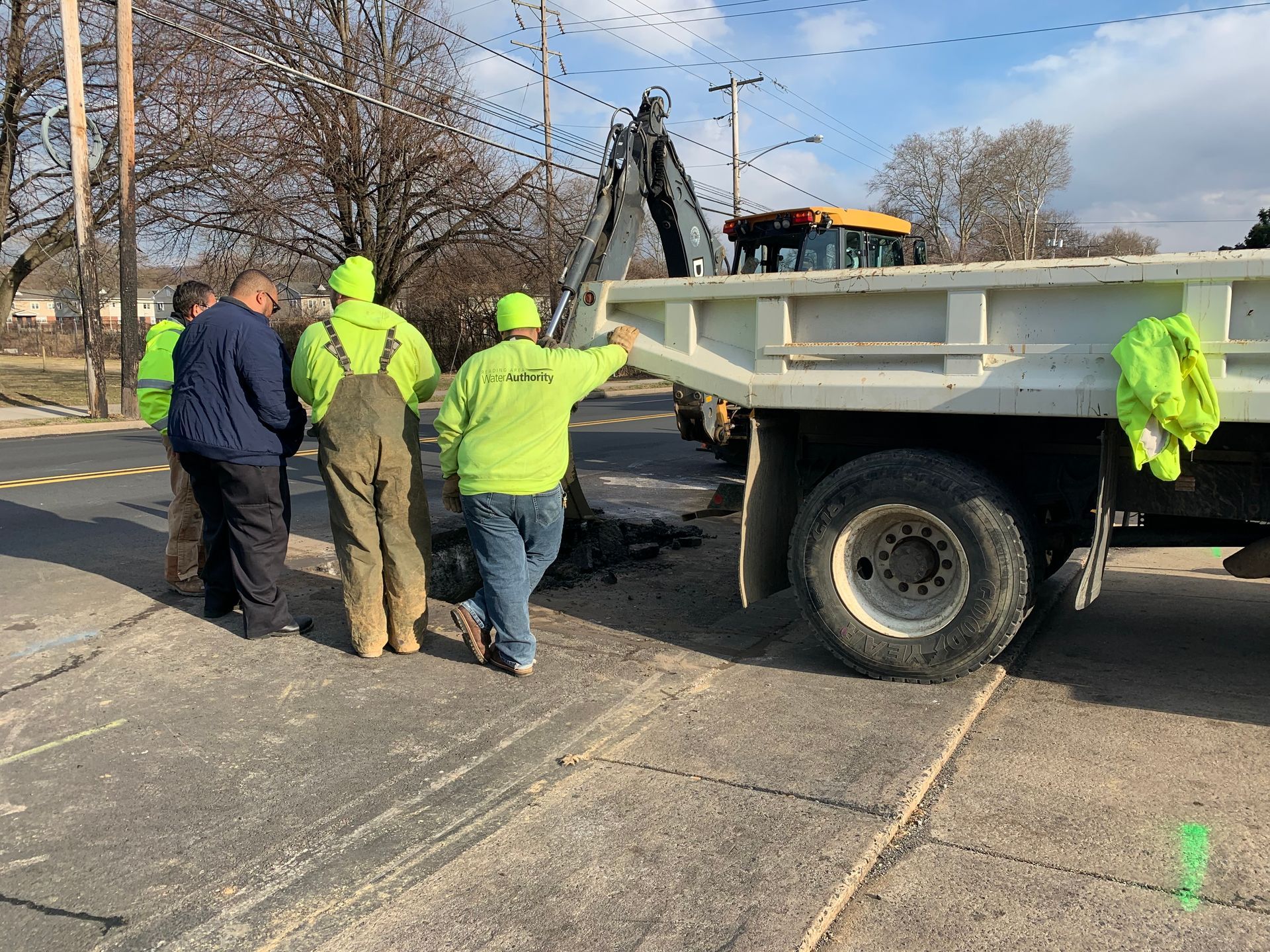 Men in safety vests watch as an excavator loads a truck with asphalt on a street.
