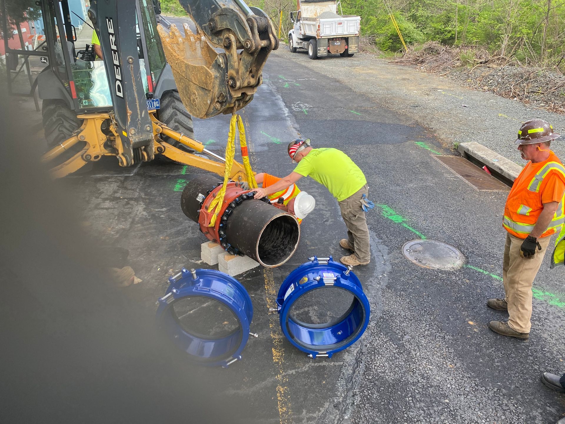 Workers installing a large pipe on a road with a backhoe. Blue rings are on the ground.