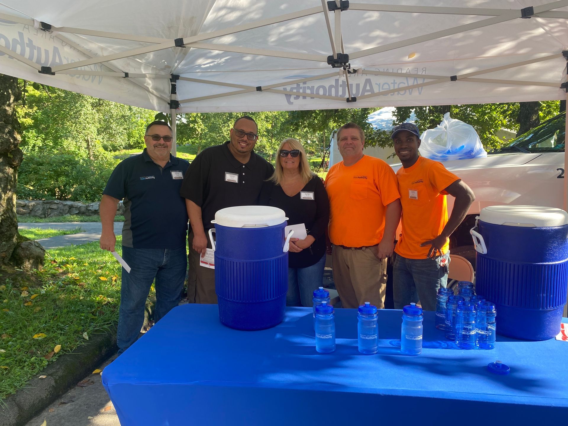 Five people stand behind a blue table at an outdoor event, next to water coolers.