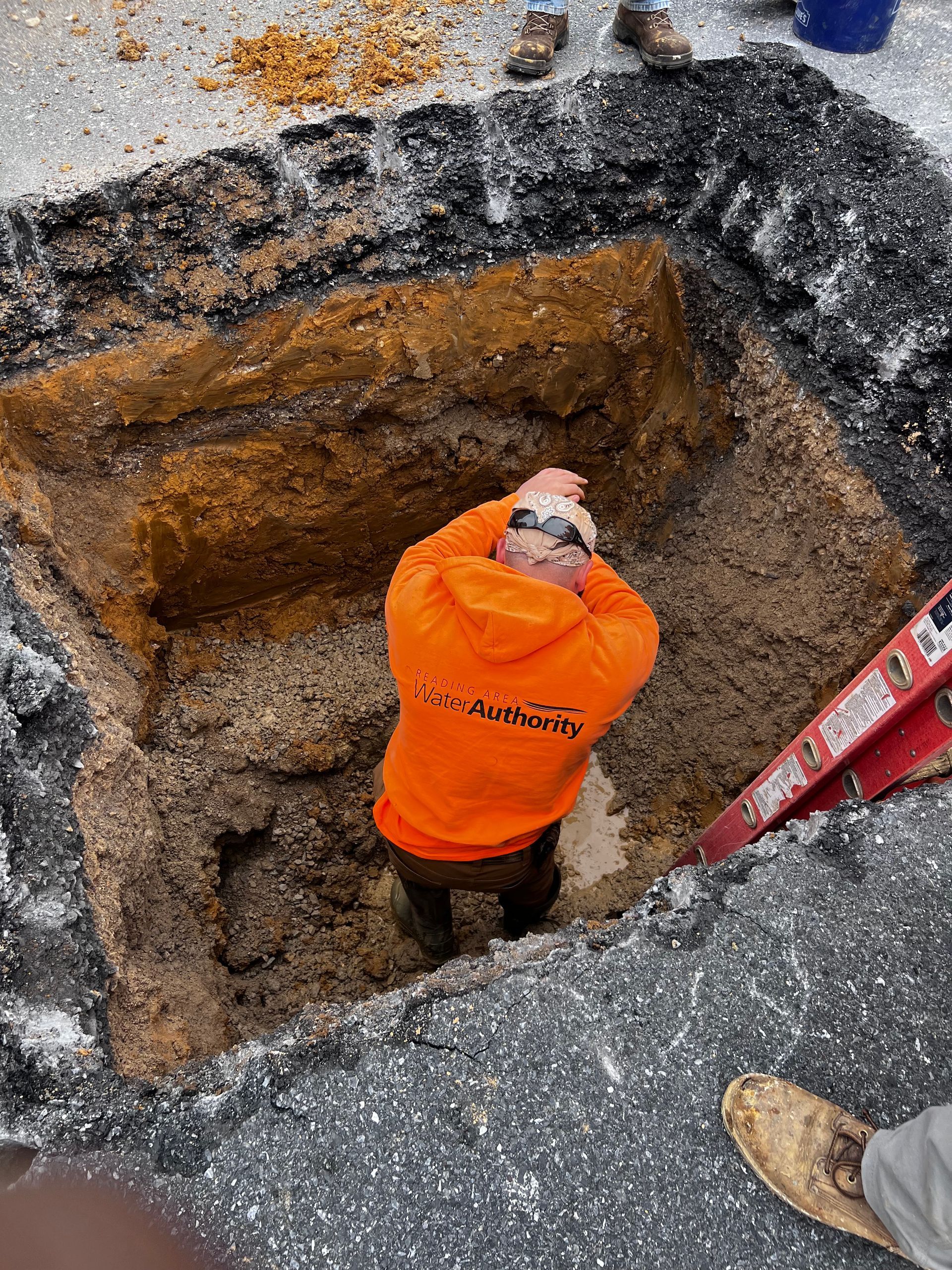 Person in orange hoodie inside a rectangular excavation in asphalt, possibly repairing underground utilities.