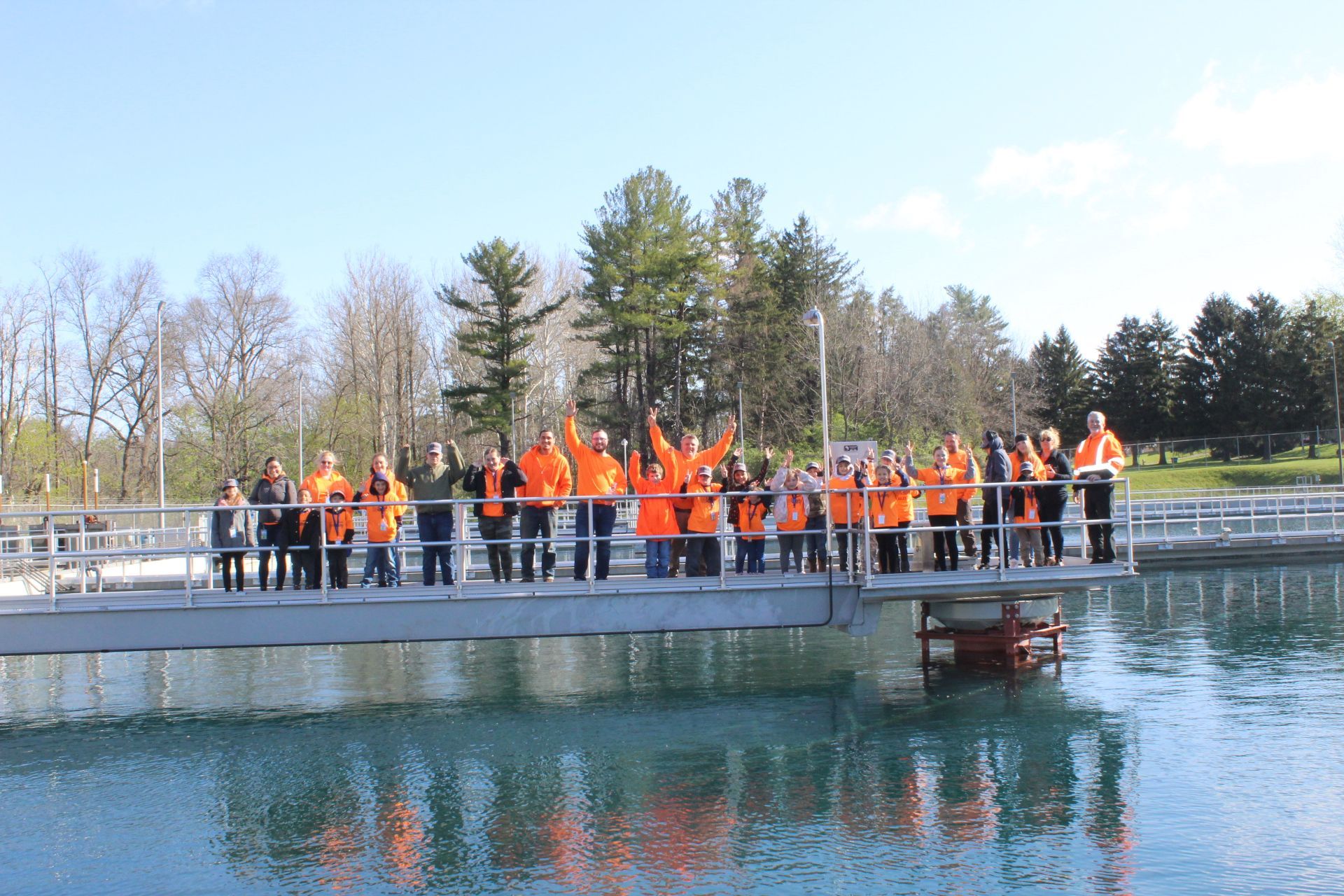 Group of people in orange vests wave from a platform over water at a water treatment facility.