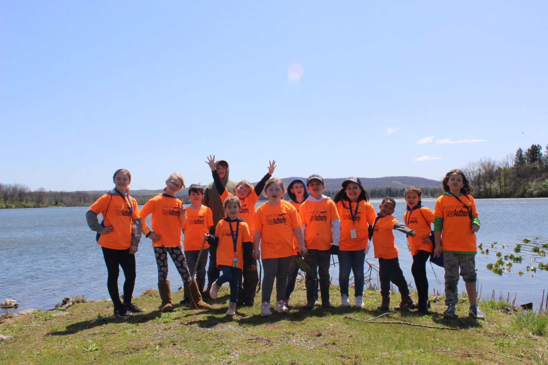 Group of children in orange shirts posing by a lake with mountains in the background under a blue sky.