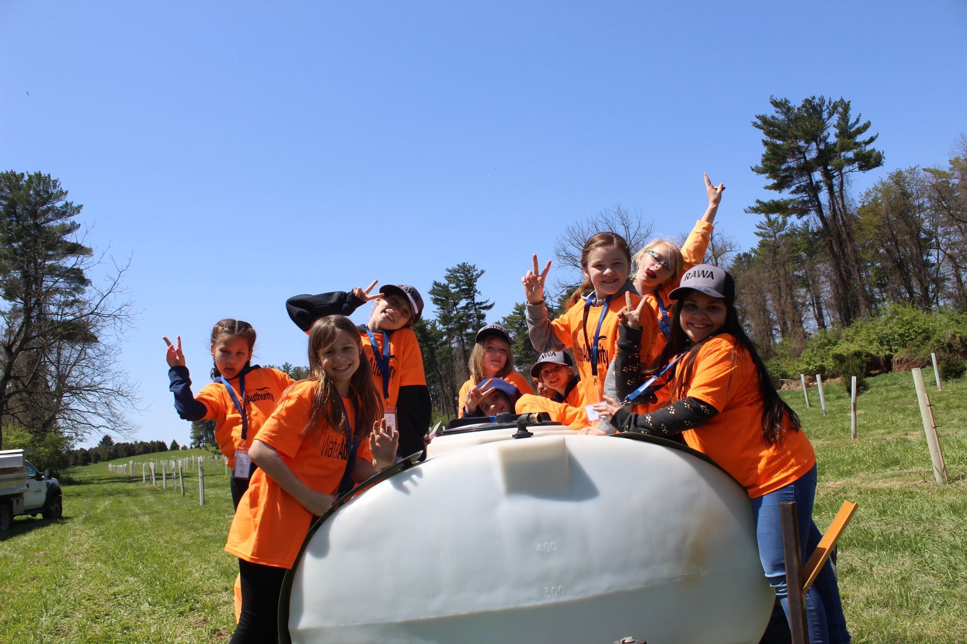 Group of children in orange shirts, smiling and posing near a large white tank outside on a sunny day.