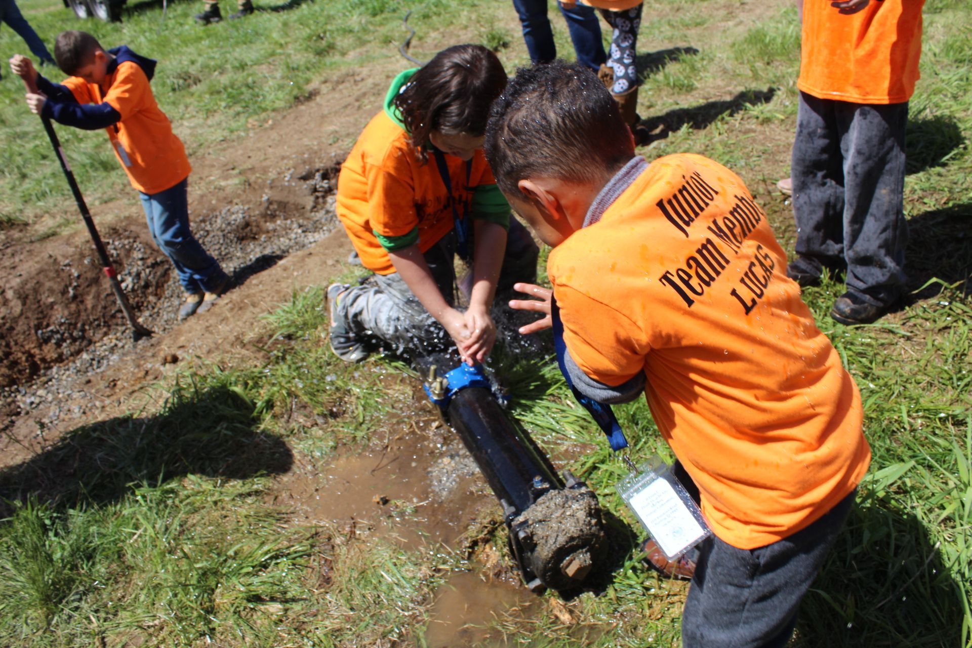 Children in orange shirts working with a black pipe outdoors, near a ditch.