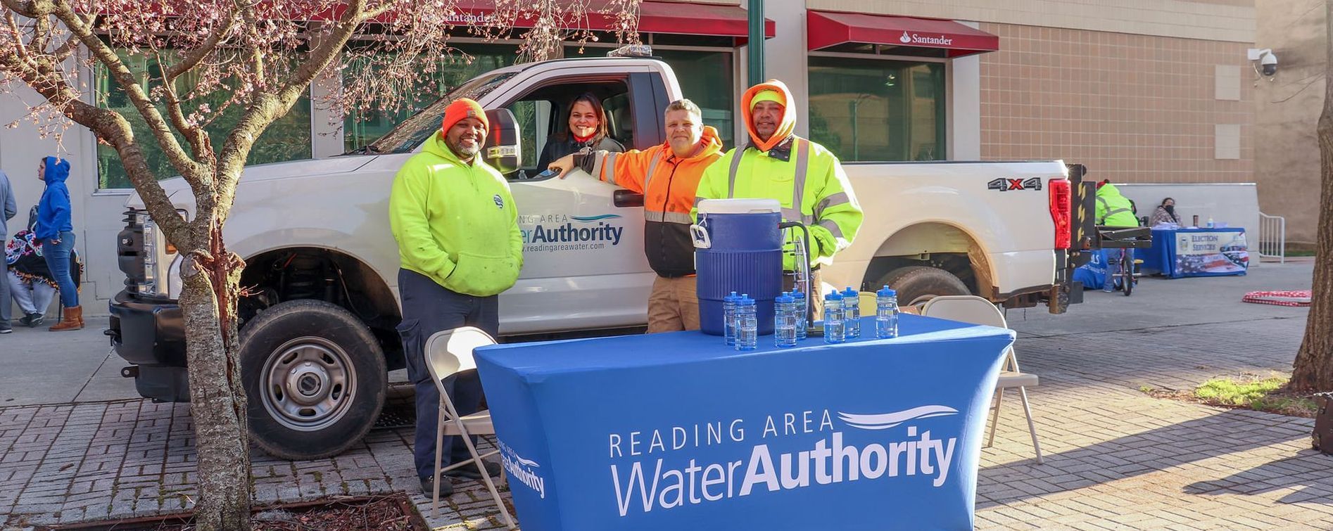 People in safety vests stand at a water authority table next to a truck, offering bottled water.