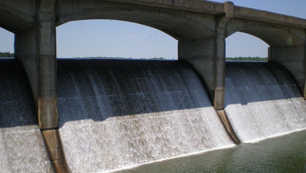 Water cascading over a dam's curved spillway, under an arched concrete structure, into a calm body of water.