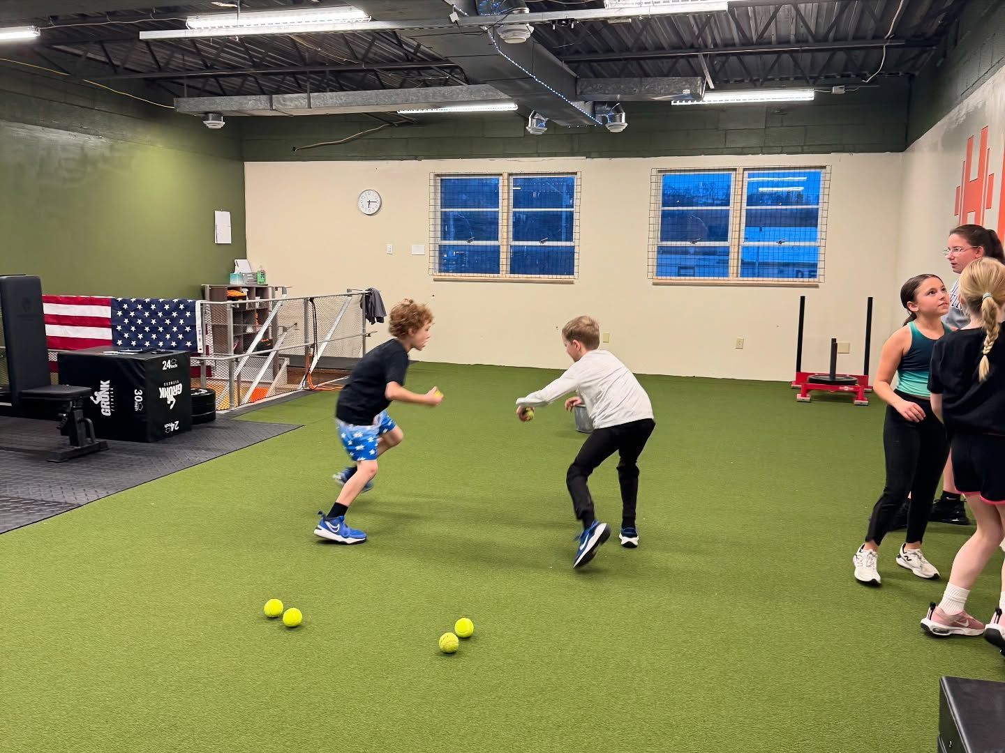 Kids playing with balls in a gym. Two boys facing each other, a coach, green turf, and an American flag.