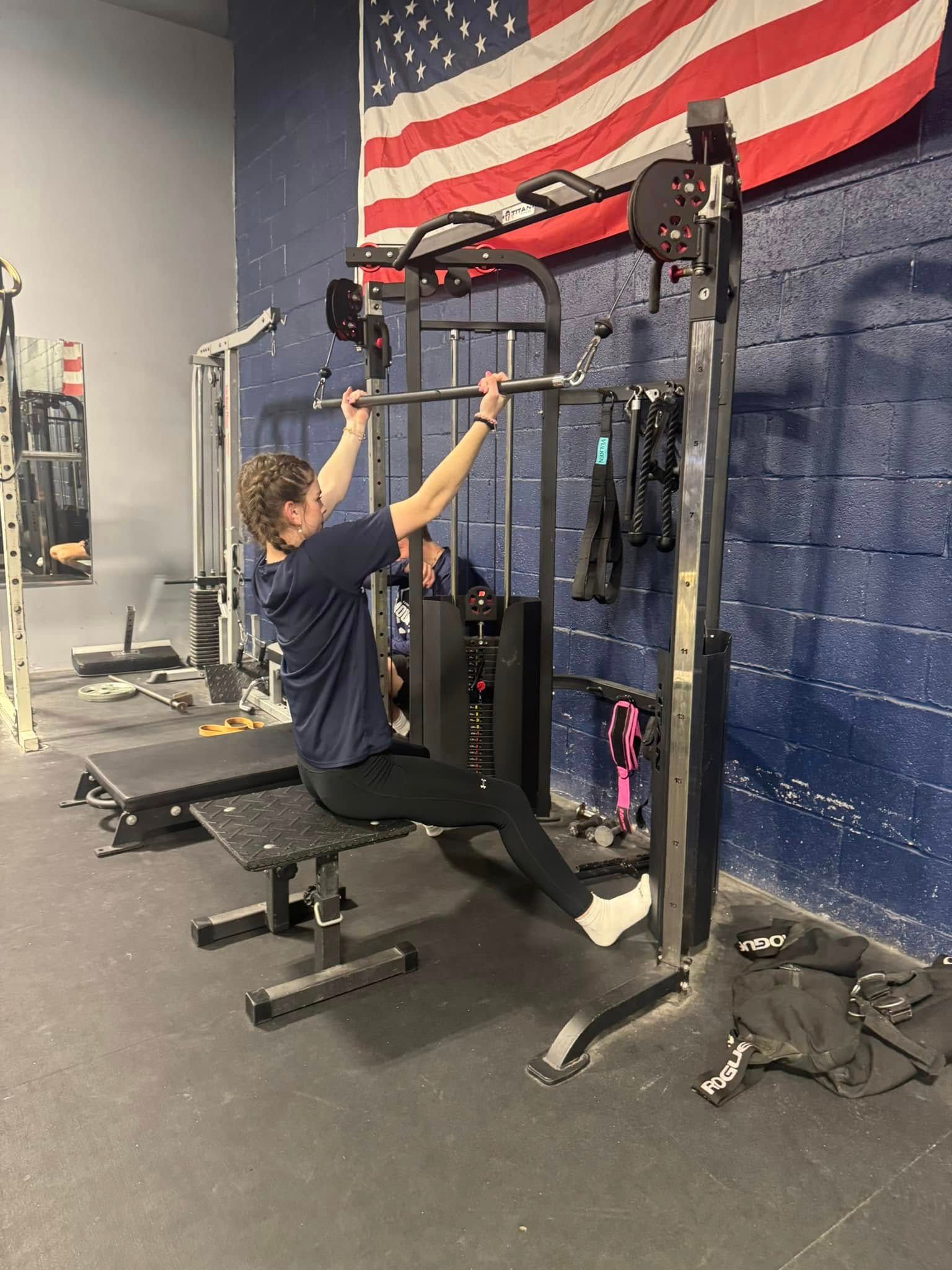 Man seated on bench, performing a pull-up using a gym machine, American flag in background.