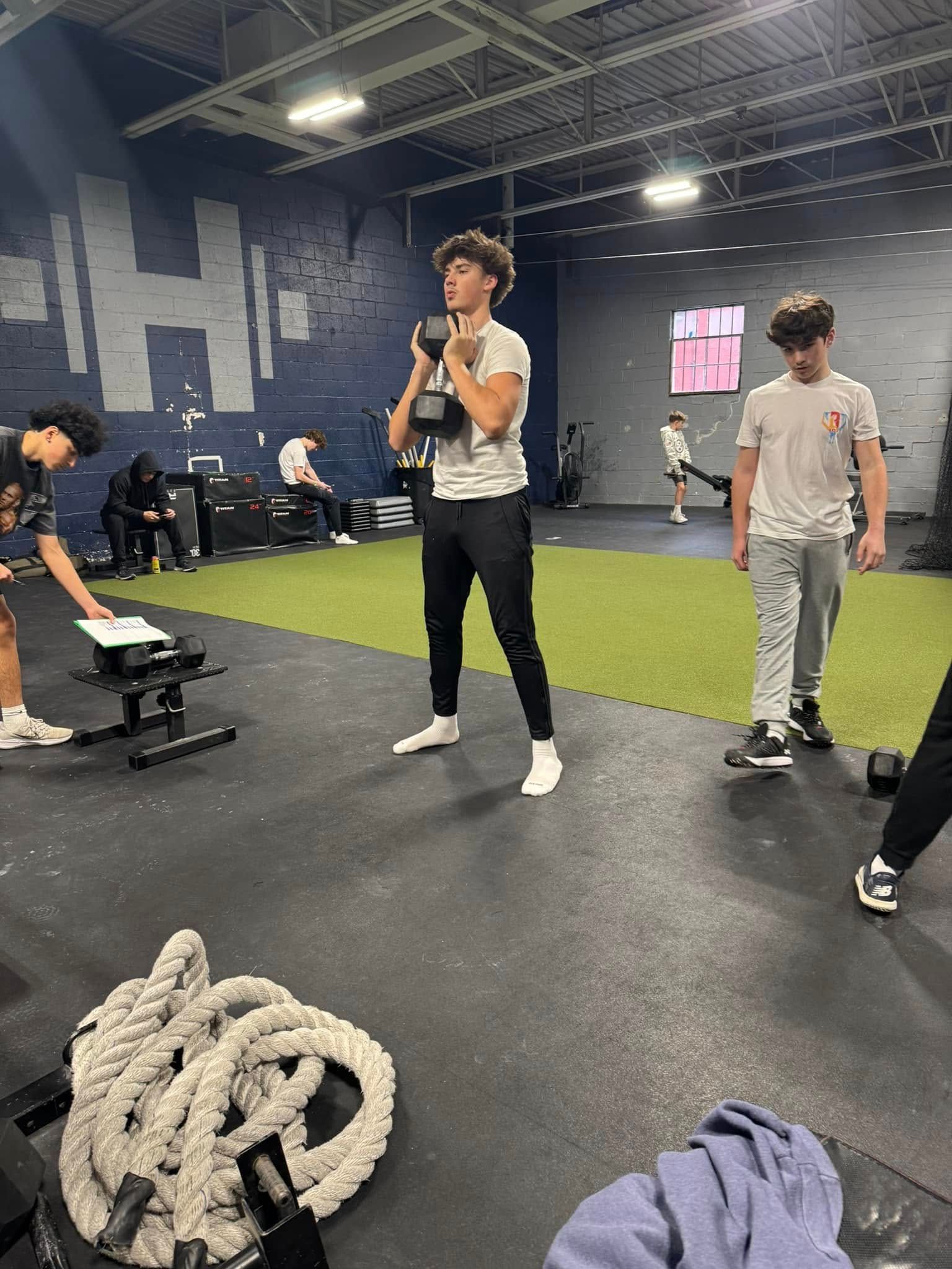 Young person holding a dumbbell in a gym; others exercise nearby. Gray floor, blue walls.