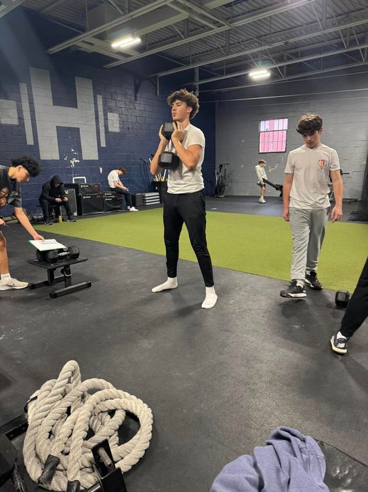Young person holding a dumbbell in a gym; others exercise nearby. Gray floor, blue walls.