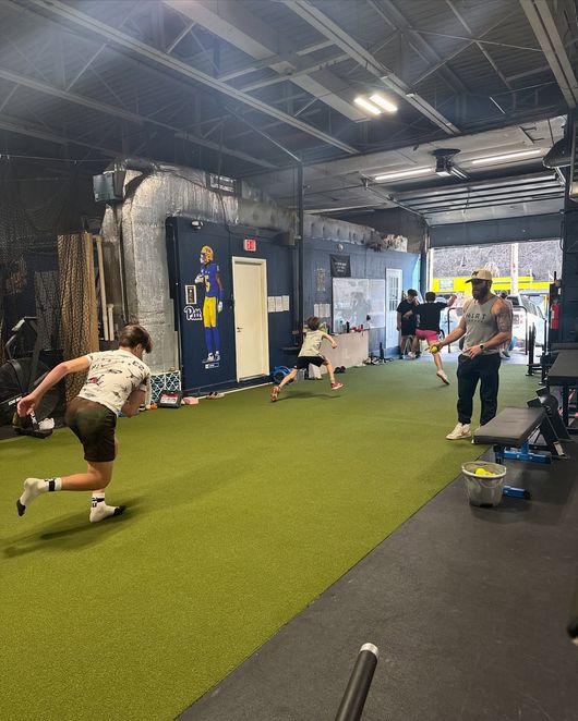 People training in an indoor gym with a coach. A person is running. Green turf on the floor.