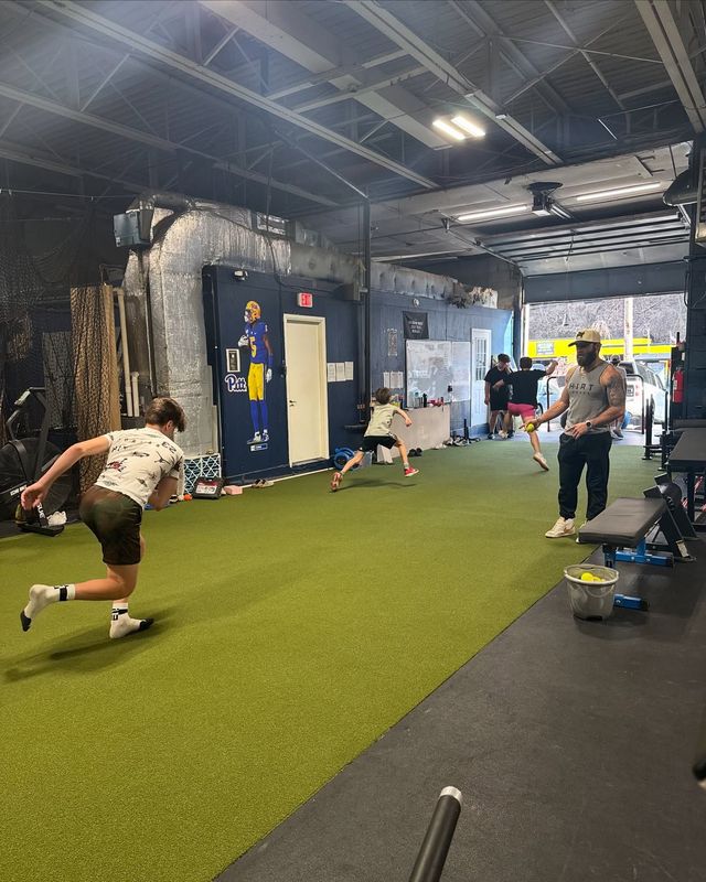 People training in an indoor gym with a coach. A person is running. Green turf on the floor.