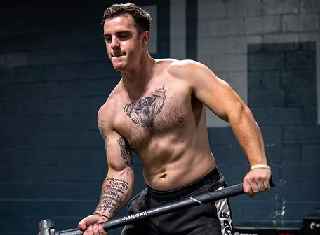 Man shirtless, exercising with a sledgehammer in a gym, chest tattoo, serious expression.
