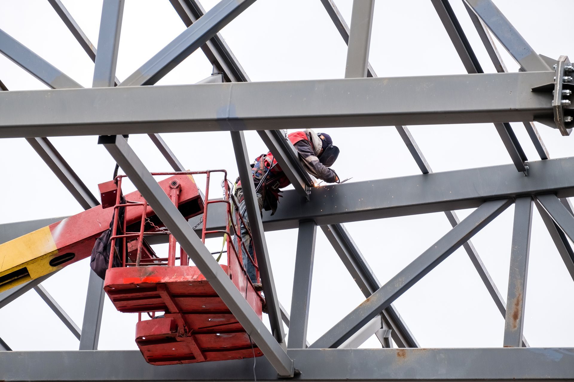 Construction worker on a lift working on steel beams of a building frame. Red lift, gray beams, overcast sky.