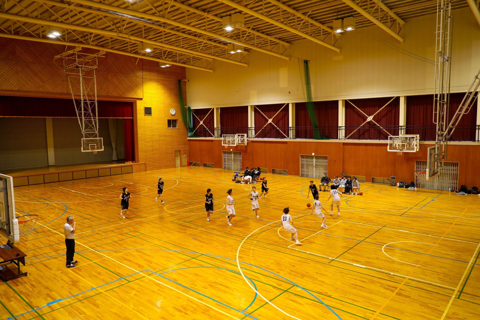 Basketball game in a wood-floored gymnasium. Players in uniforms are on the court. Referee watches.