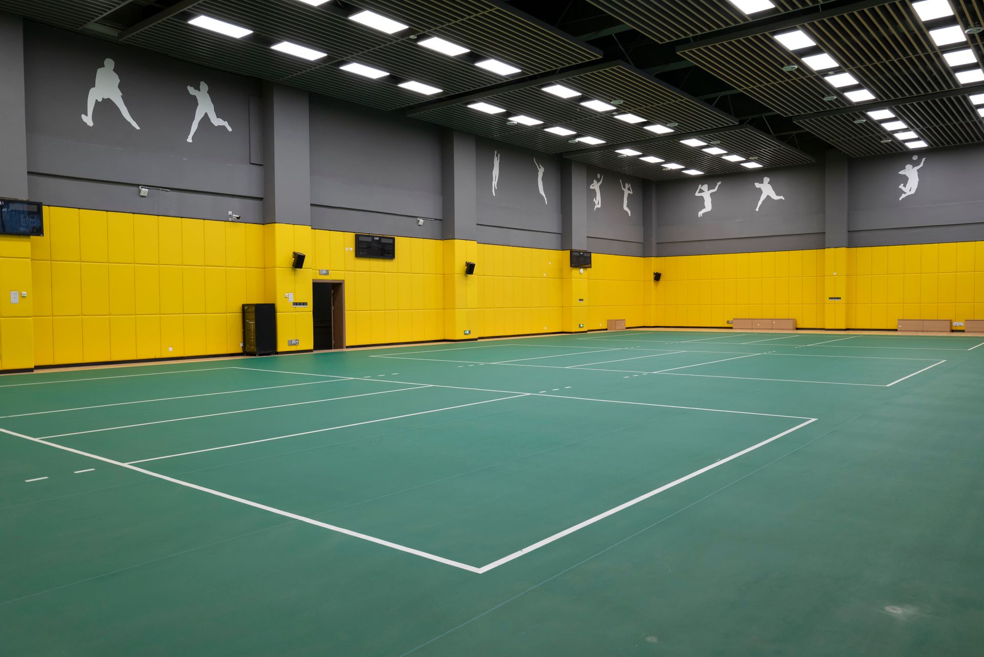 Indoor sports court with green floor, white lines, yellow wall padding, and silhouettes of athletes.