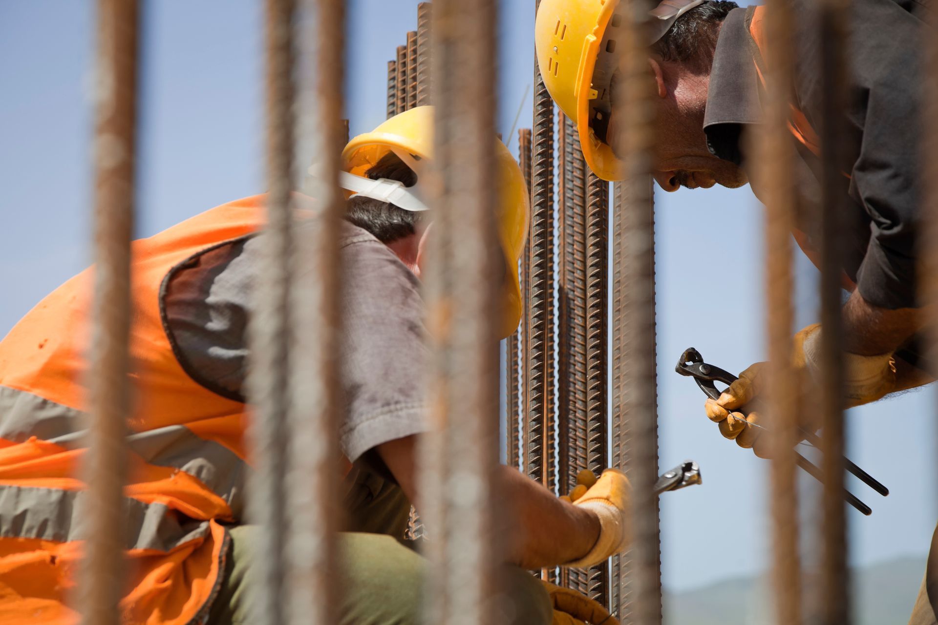 Two construction workers in yellow helmets and orange vests, securing rebar.