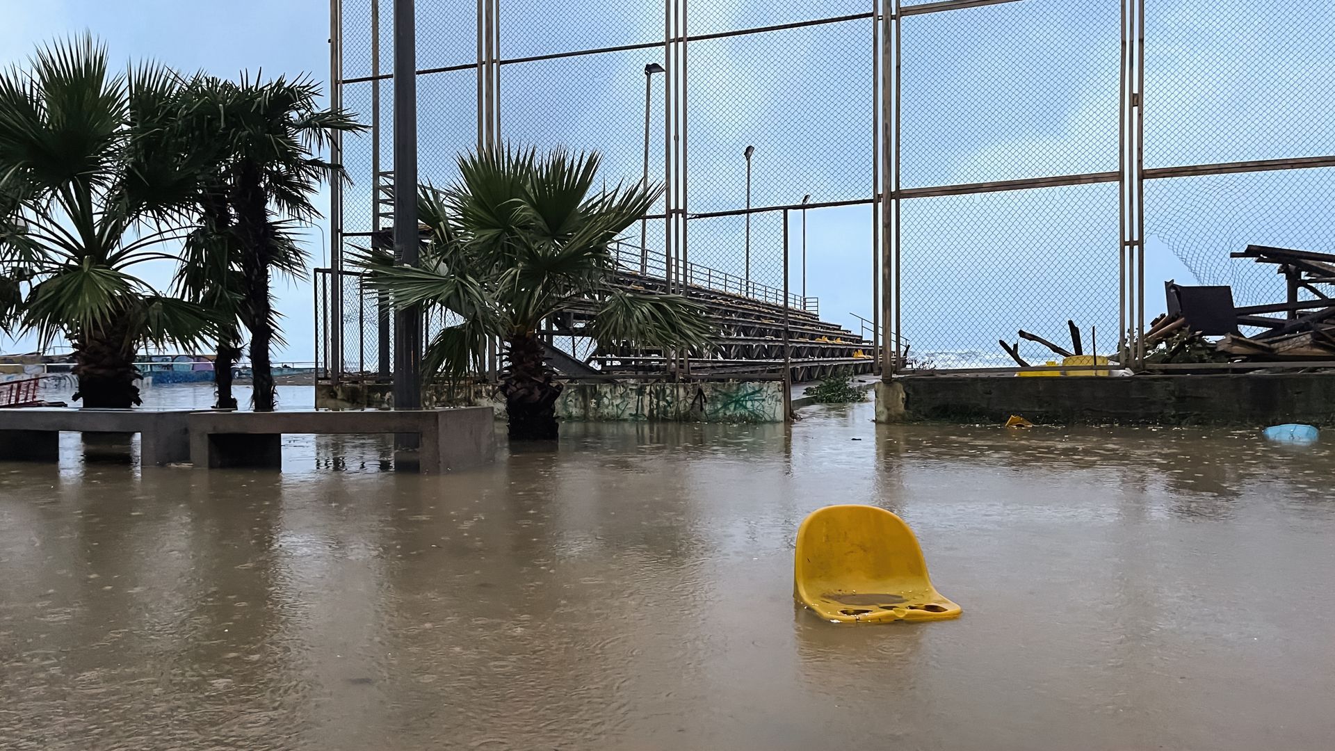 Floodwater engulfs a waterfront area with a yellow chair floating. Palm trees and a fence are visible.