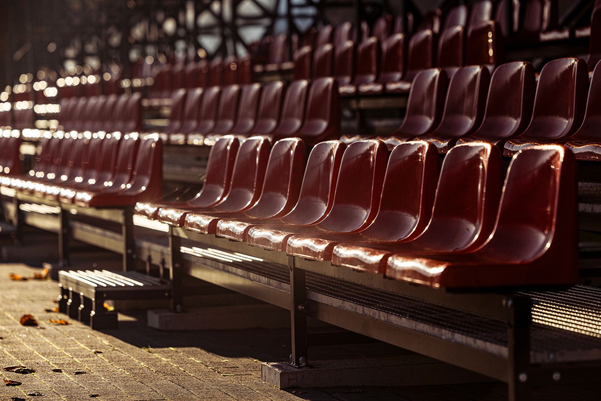 Rows of empty red stadium seats, brightly lit, with shadows on the ground.