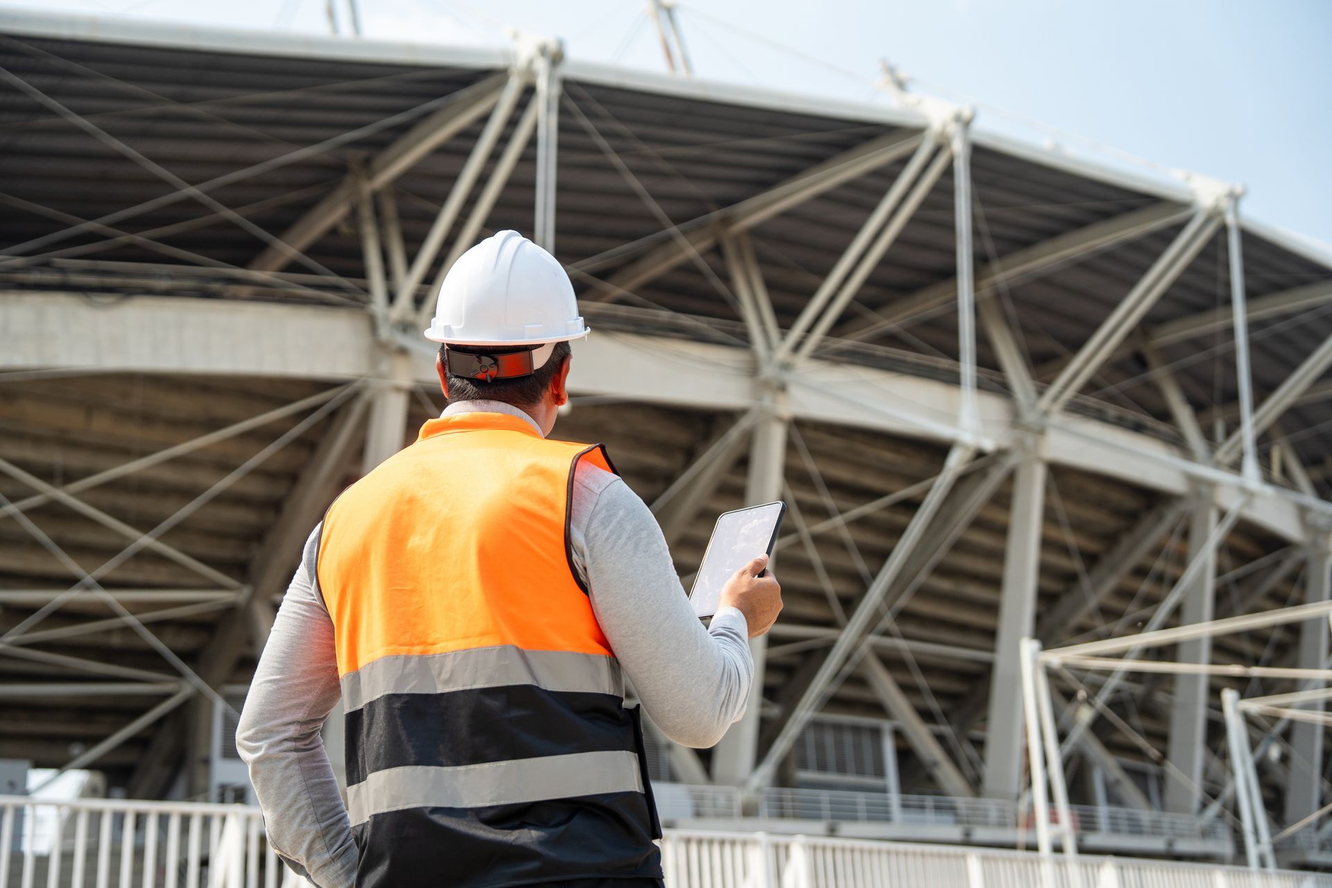 Construction worker in white hard hat and orange vest, reviewing blueprints at a stadium.