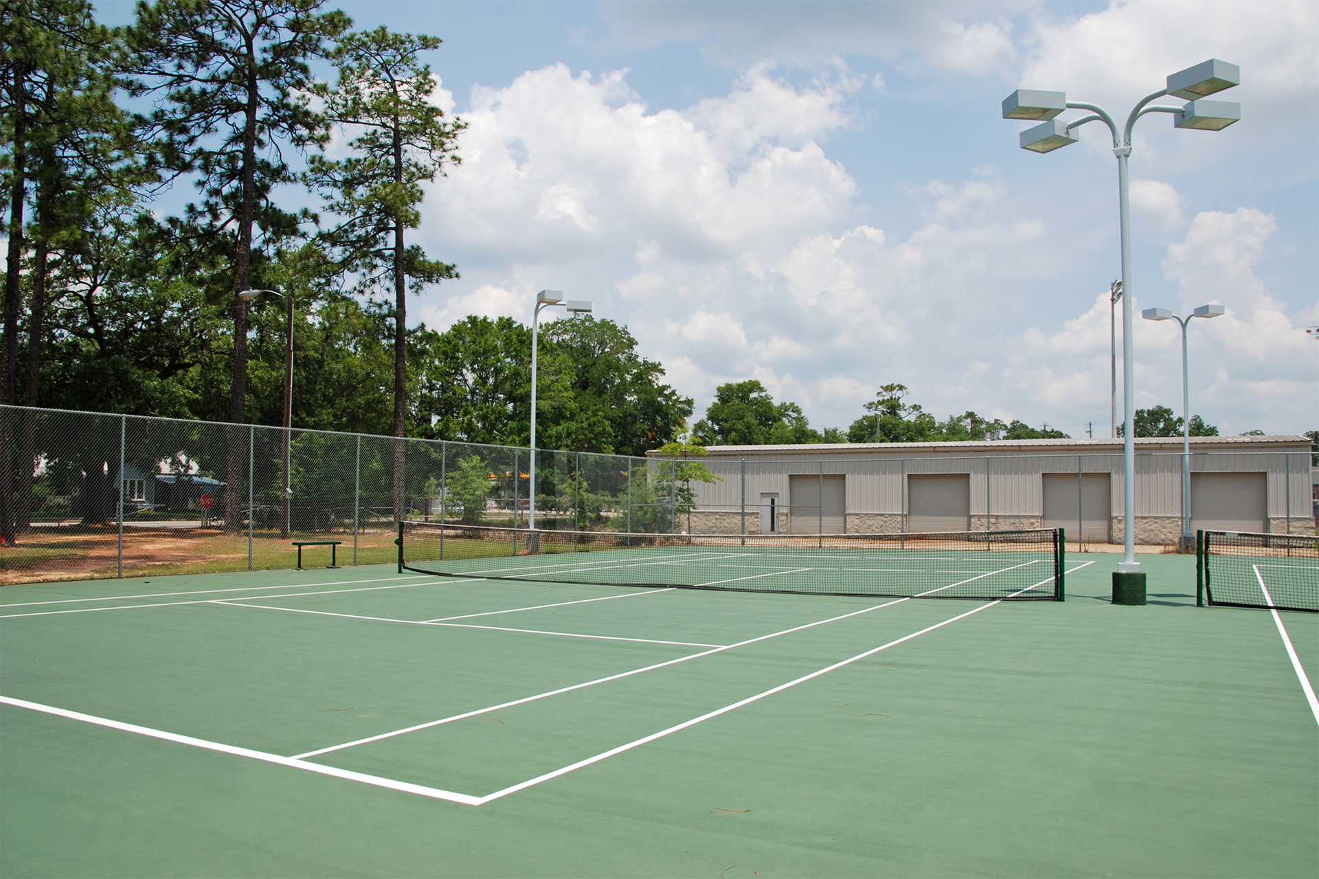 Tennis court with green surface, white lines, net, and tall light poles, under a partly cloudy sky.