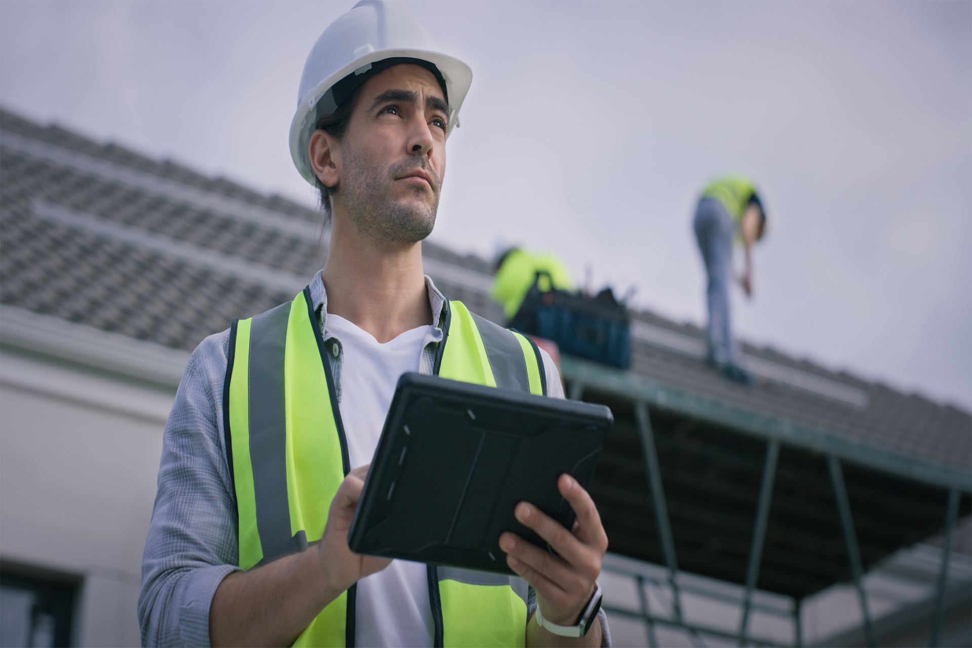 Construction worker in hardhat and vest using a tablet, looking up at workers on a roof.