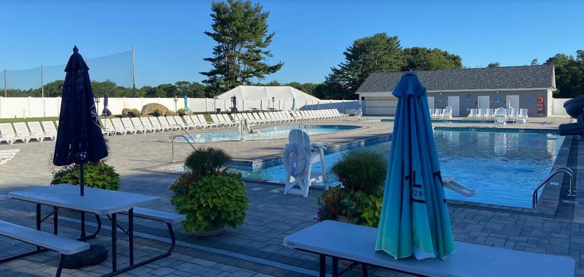 A swimming pool with umbrellas and picnic tables in front of it.