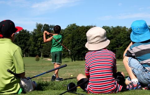 A group of young boys are sitting on the grass watching a boy swing a golf club.