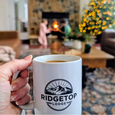 Hand holding white mug with Ridgetop Lodge logo by fireplace.
