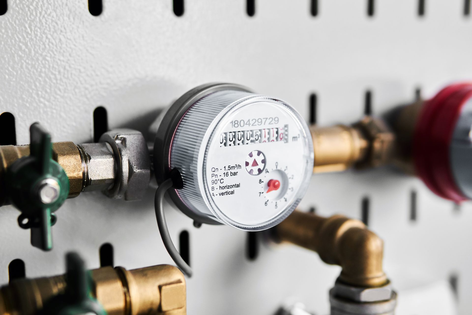 A water meter is hanging on a wall next to brass pipes.
