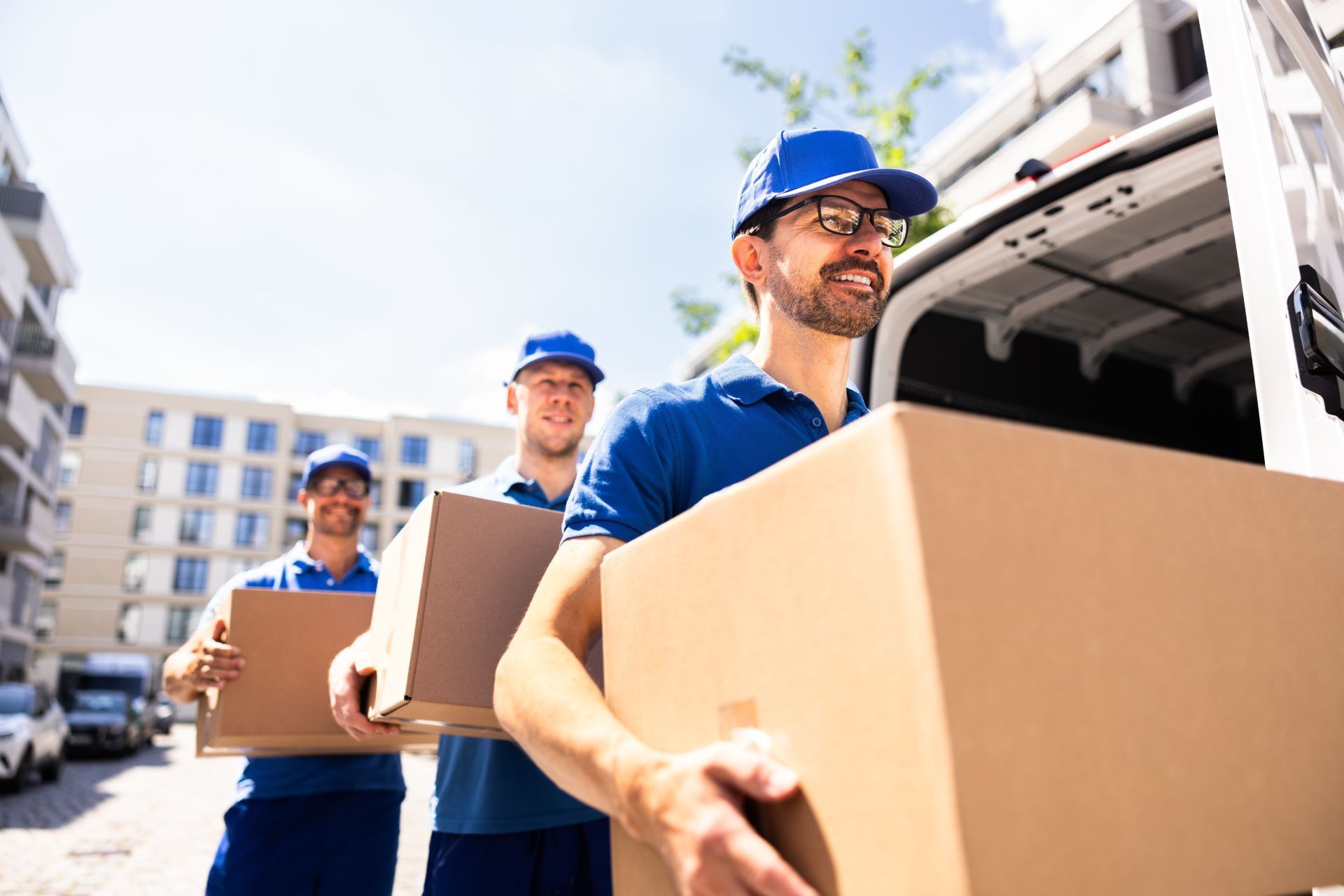 Movers in blue uniforms carry cardboard boxes from a van outdoors.