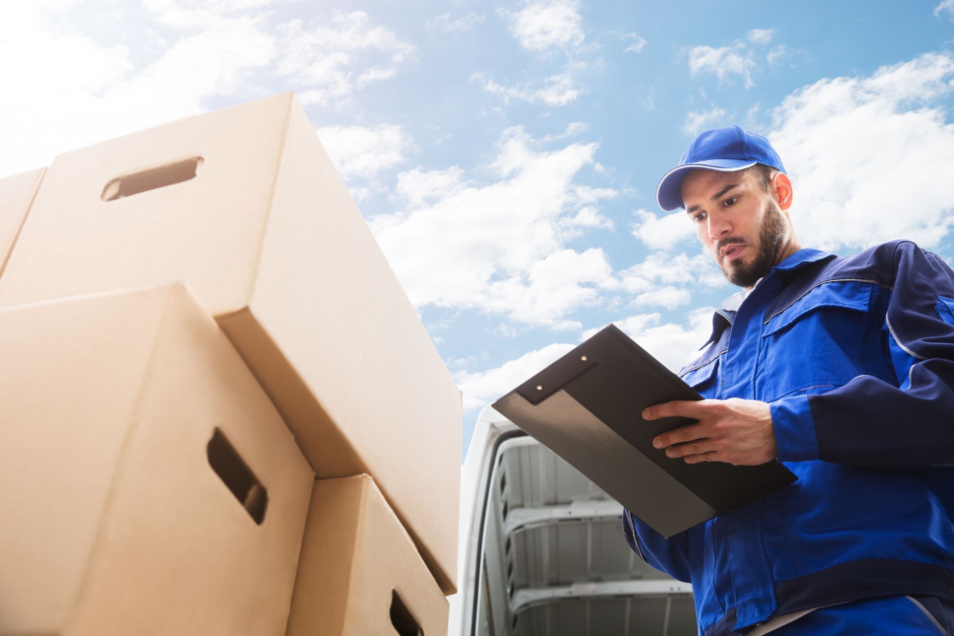Delivery worker in blue uniform, looking at clipboard near stacked cardboard boxes outside.
