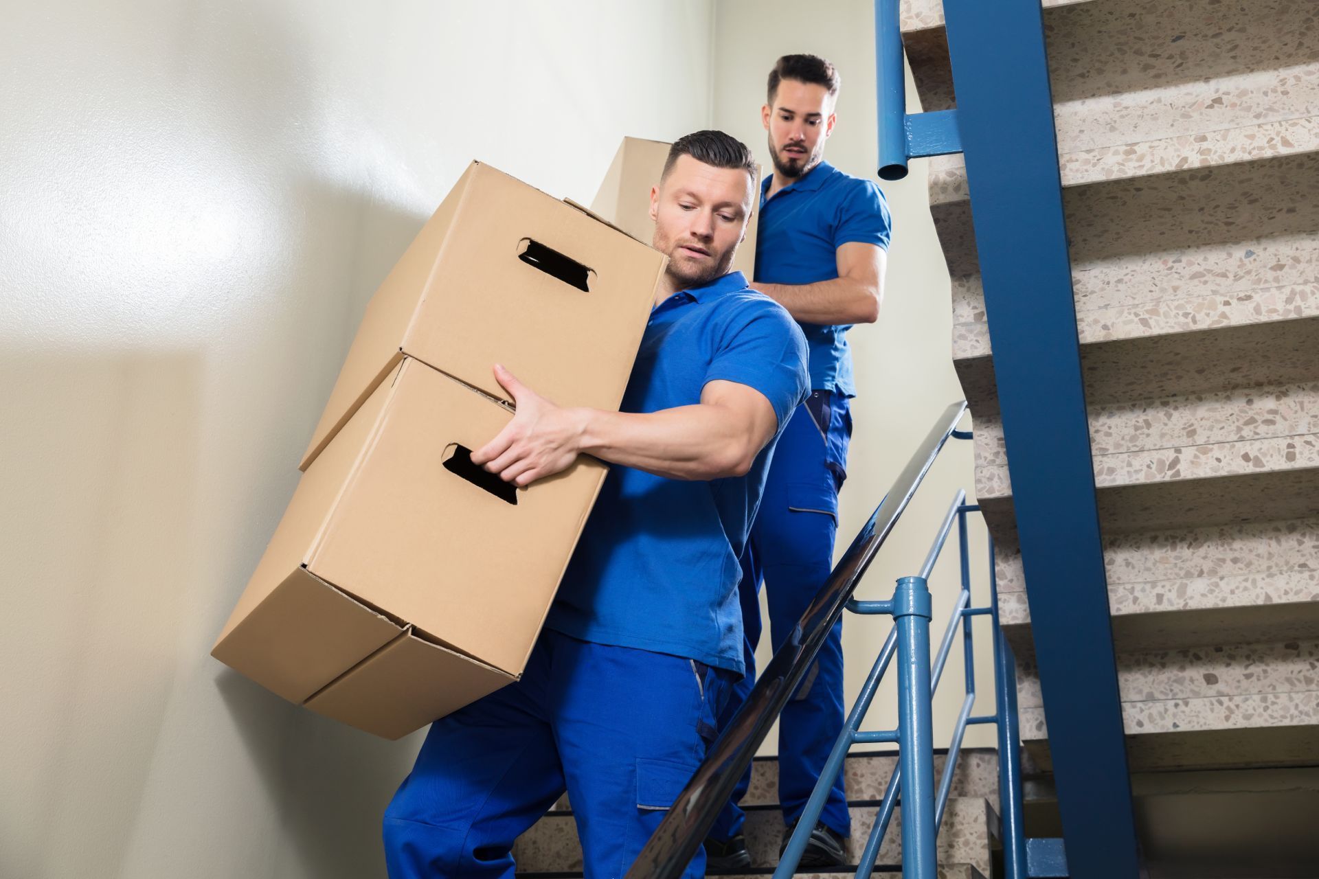 Two movers in blue uniforms carrying stacked boxes up a staircase.