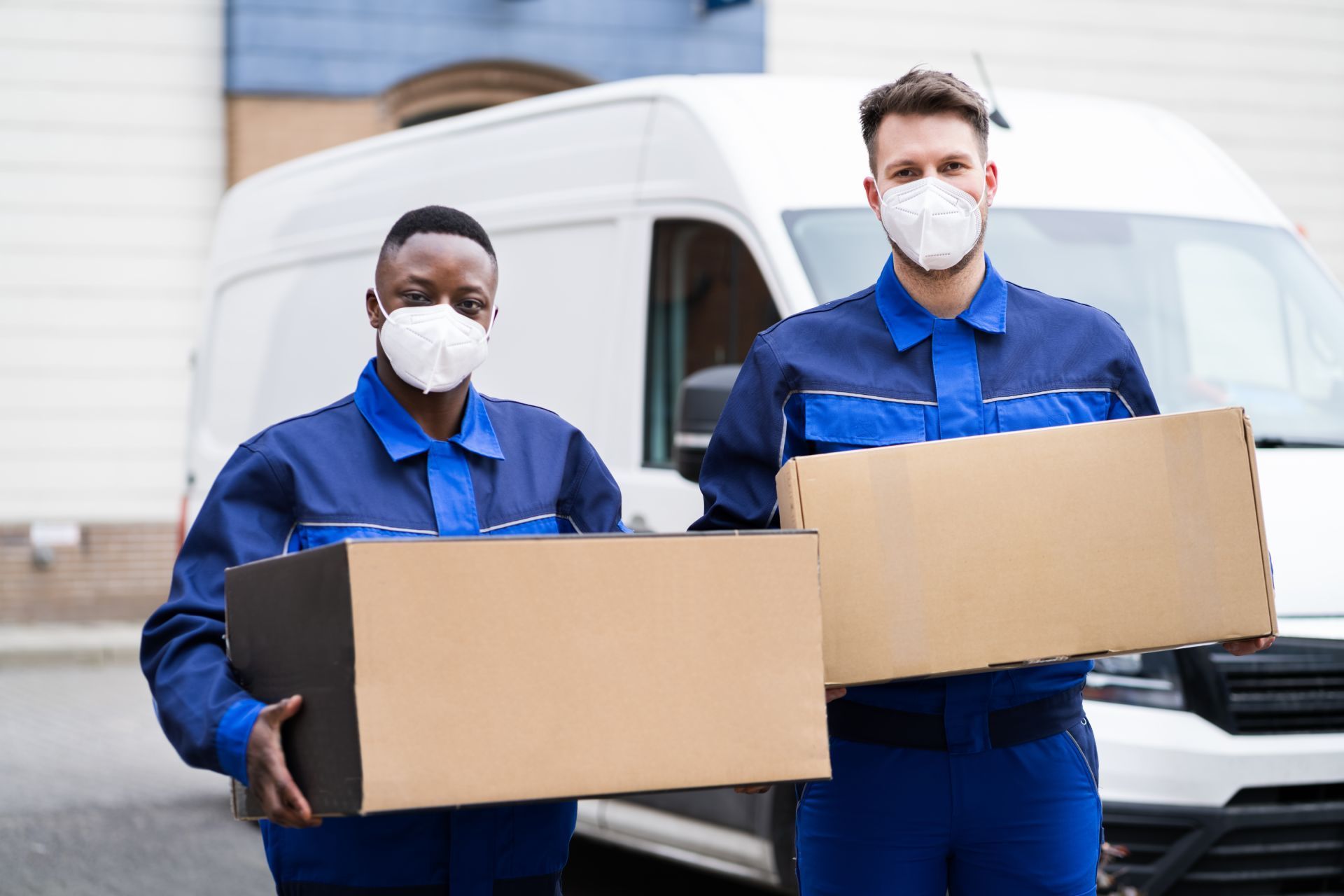 Two people in blue jumpsuits, wearing masks, carrying boxes in front of a white van.