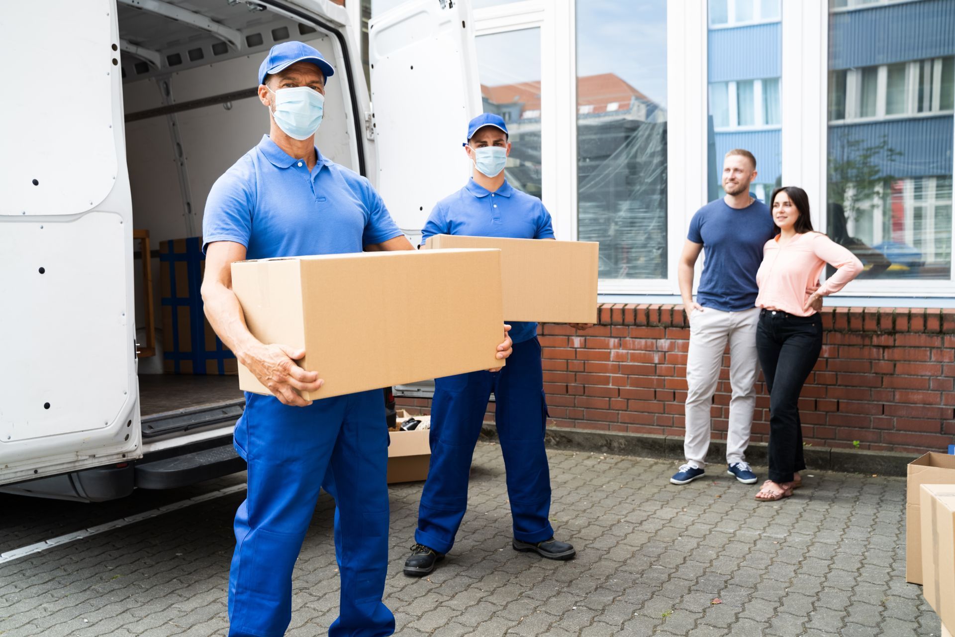 Movers in blue uniforms carrying boxes, van in background, couple watching.