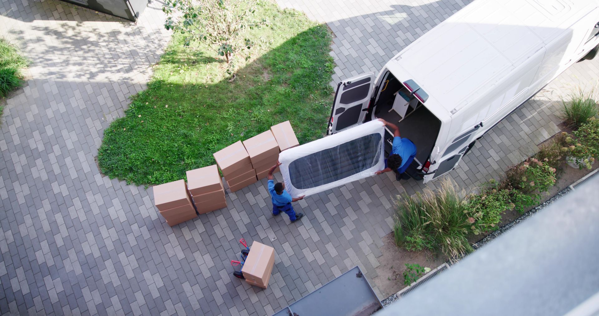 Movers loading a mattress and boxes into a white van parked outside a building on a brick path.