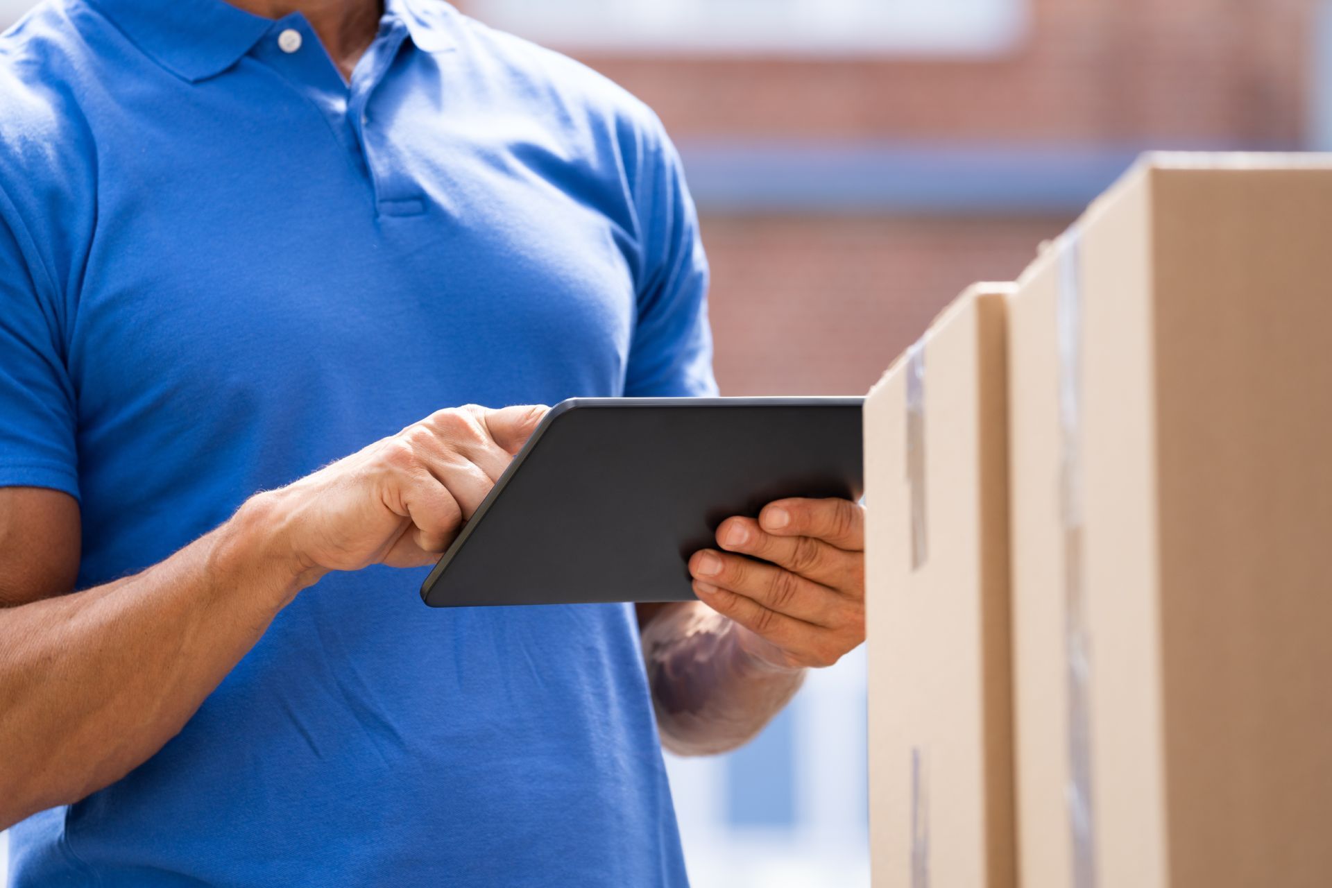 Delivery person in blue polo shirt using tablet near cardboard boxes outside.