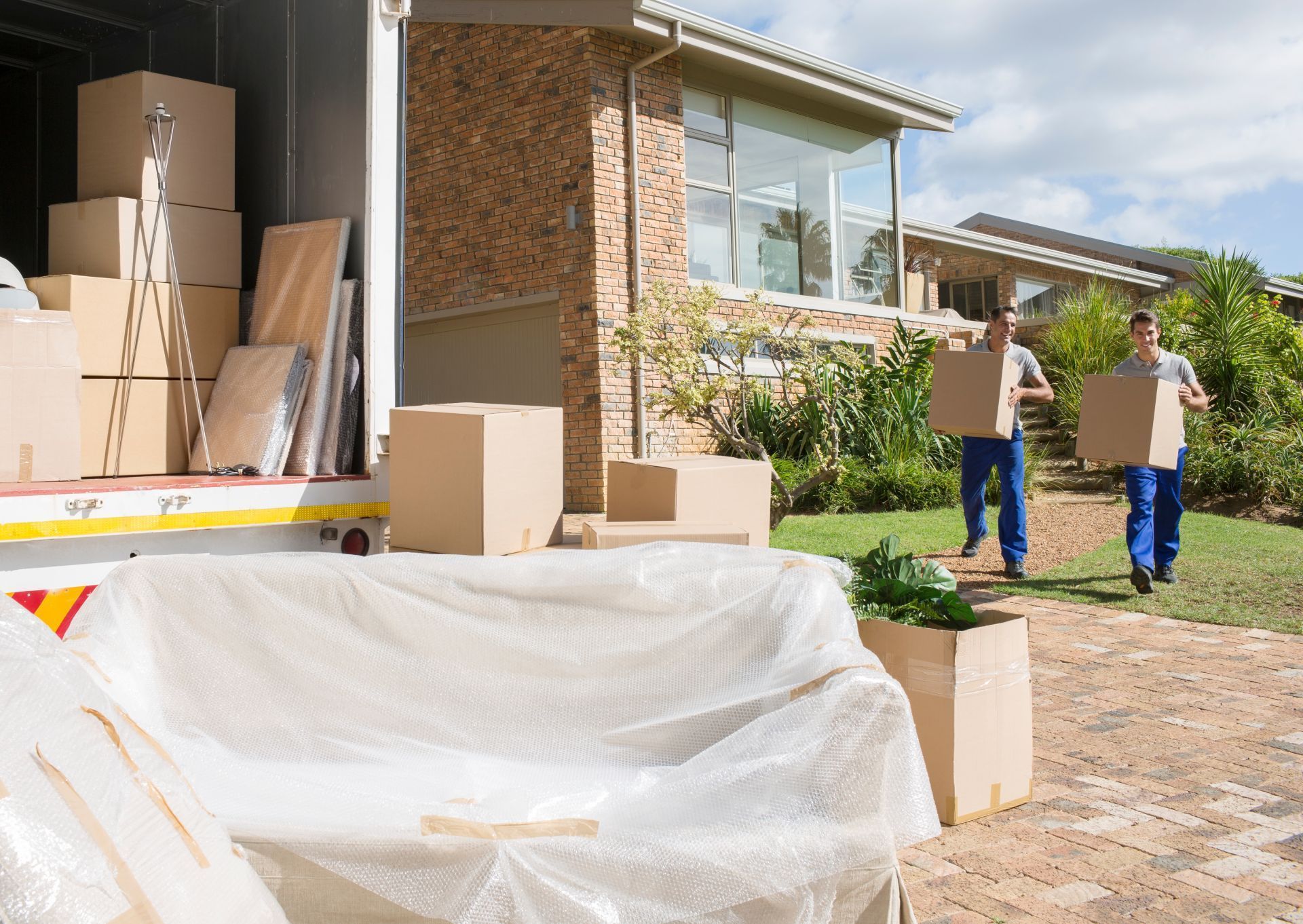 Two movers carrying boxes from a moving truck to a house. Furniture wrapped in bubble wrap.