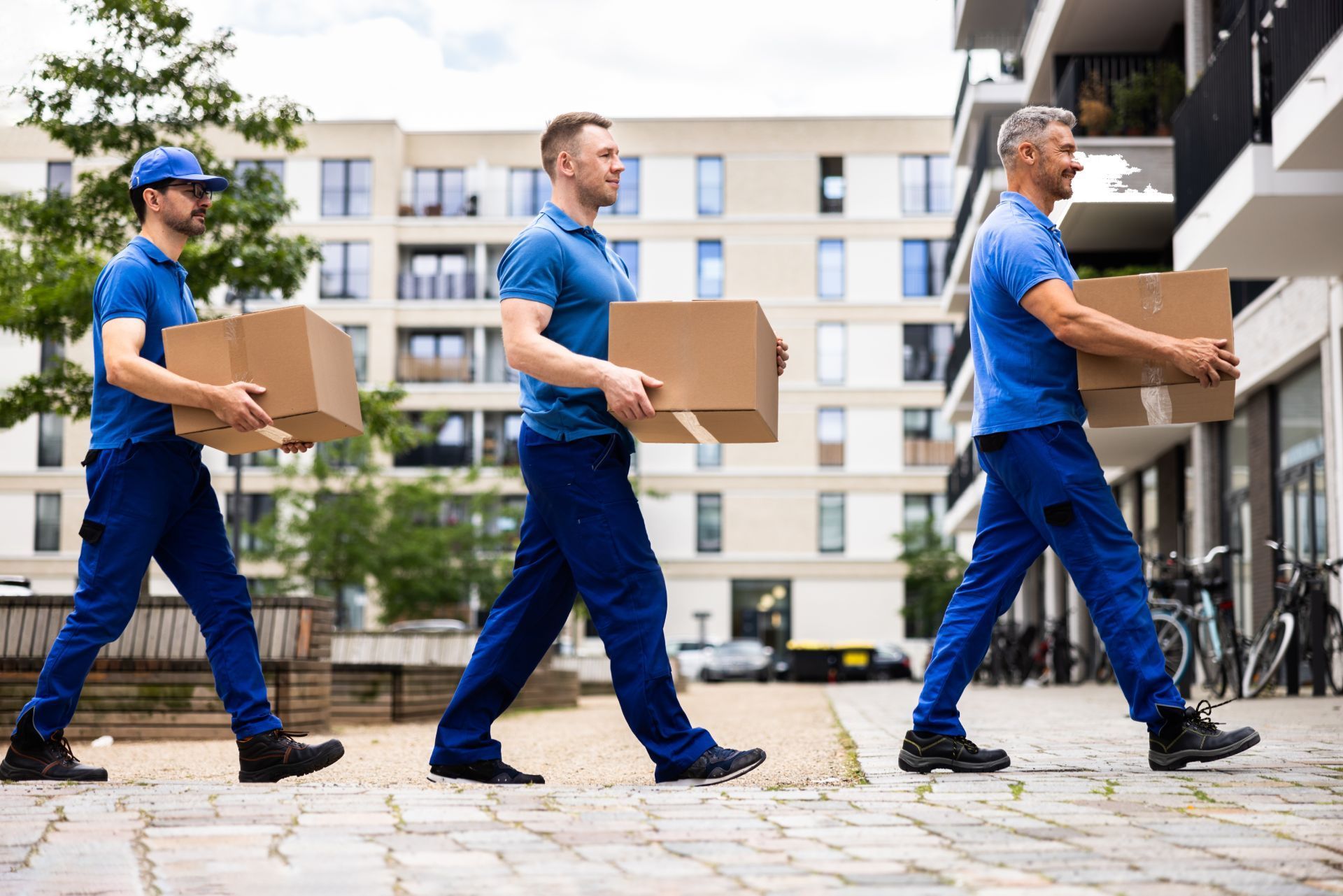Three movers in blue uniforms carrying boxes on a city street.