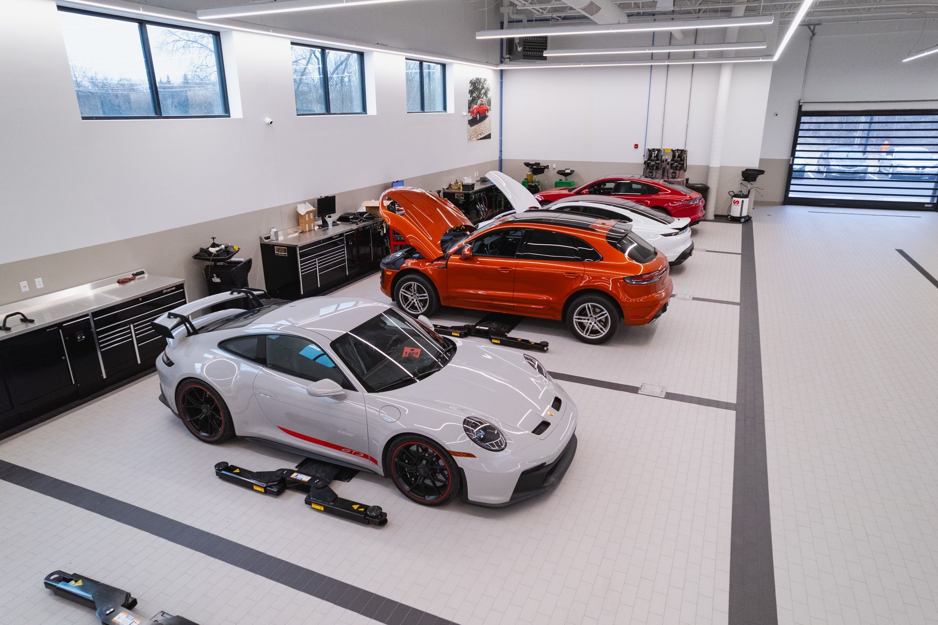 A high-angle view of a clean, bright auto repair garage featuring a gray Porsche, an orange SUV, and other parked cars.