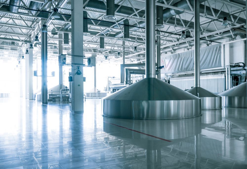 Large, shiny stainless-steel industrial vats in a bright, spacious factory with a reflective floor and exposed ceiling.
