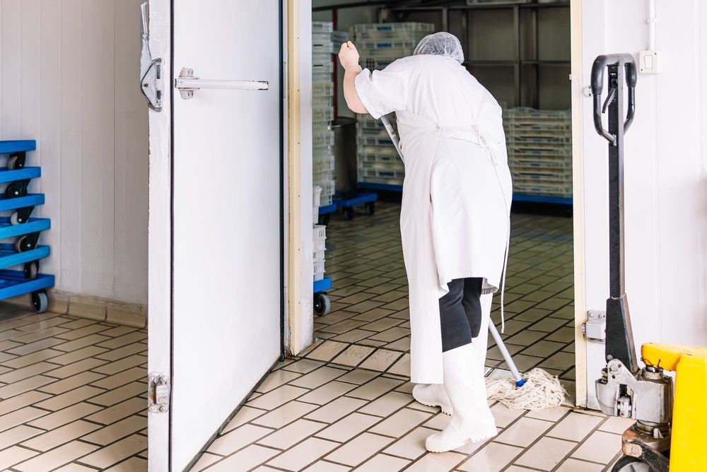 A person in protective white clothing mops the tiled floor of a walk-in industrial freezer.