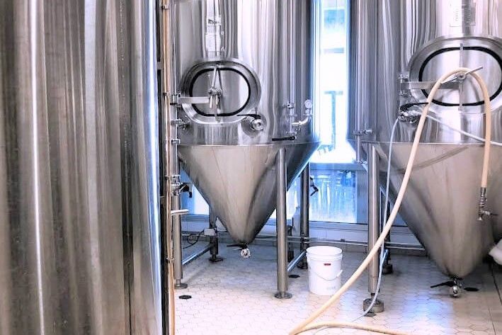 Large stainless steel fermentation tanks in a brewery with a white bucket on the floor.