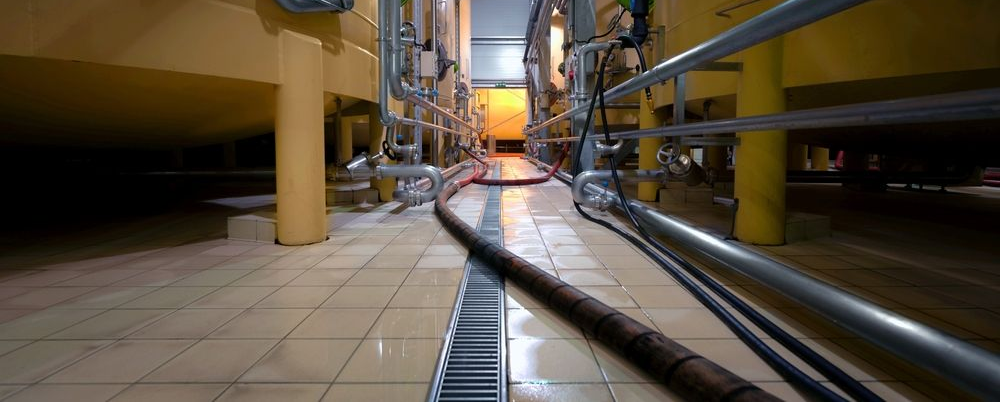 A low-angle view of a tiled aisle between two large, yellow industrial tanks connected by metal pipes and conduits.