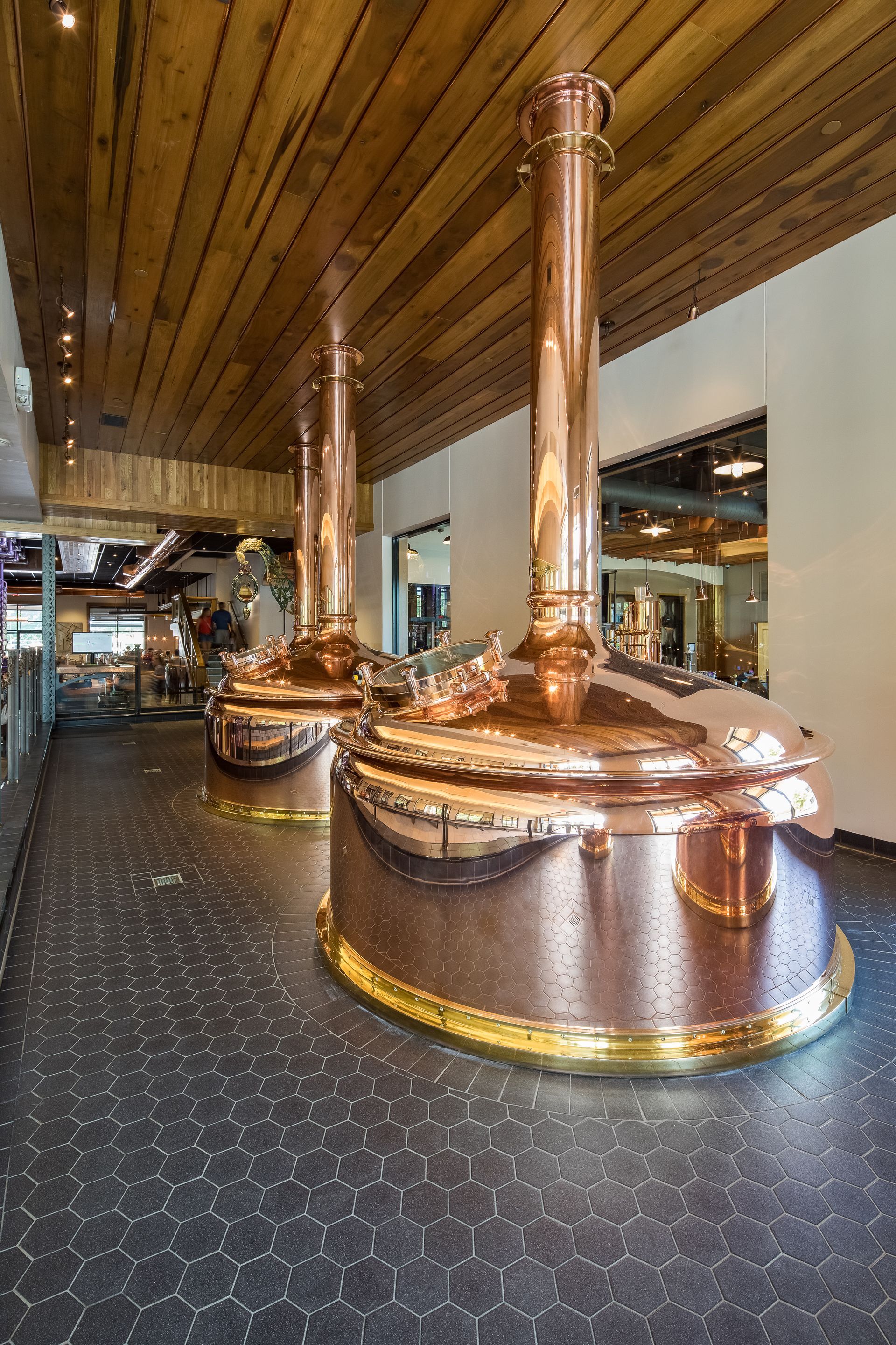 Two copper brewing tanks in a brewery. Wooden ceiling, hexagonal black tiled floor, and a mirror are also visible.